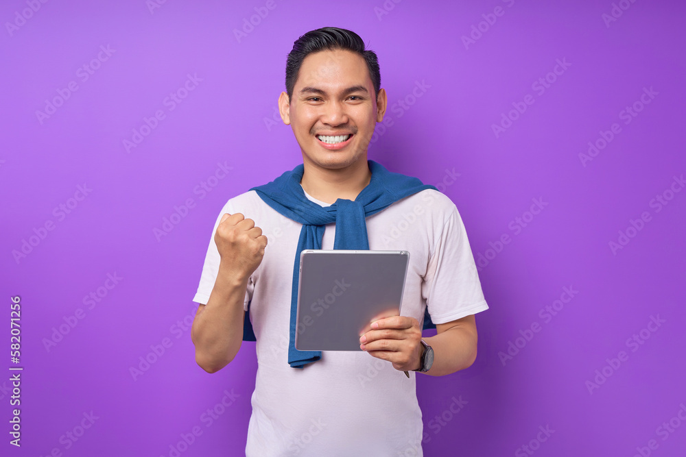 Excited young Asian man in t shirt holding digital tablet and rejoicing winning online with excitement isolated over purple background Excited young Asian man in t shirt holding digital tablet and rejoicing winning online with excitement isolated over purple background