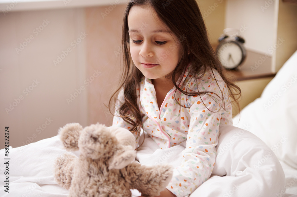 Adorable elementary age Caucasian child girl in stylish pajamas, sitting on bed after waking up in the morning, playing with a plush toy sheep, in her cozy bedroom. Children. Happy childhood concept Adorable elementary age Caucasian child girl in stylish pajamas, sitting on bed after waking up in the morning, playing with a plush toy sheep, in her cozy bedroom. Children. Happy childhood concept