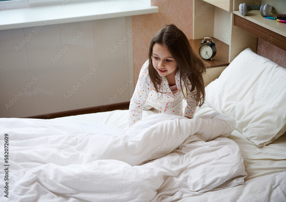 Lovely happy Caucasian child girl in stylish white pajamas with colorful dots, waking up in a happy mood, smiling while and folding her bed and cleaning her bedroom in the sunny morning Lovely happy Caucasian child girl in stylish white pajamas with colorful dots, waking up in a happy mood, smiling while and folding her bed and cleaning her bedroom in the sunny morning