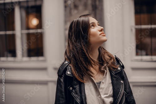 Trendy girl with brunette hairstyle posing outside. Woman in leather jacket and shirt look at size walking on street outdoors. Tourist happy woman posing in the city. Optimistic lady walks outside.