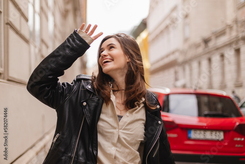 Trendy girl with brunette hairstyle posing outside. Woman in leather jacket and shirt walking on street outdoors. Tourist happy woman posing in the city. Optimistic lady walks outside, hand near face.