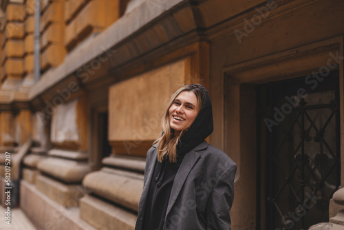 Trendy girl with blonde hairstyle posing outside. Woman in grey jacket and hoodie walking on the street outdoors. Tourist happy woman posing in the city. Optimistic lady walks outside.