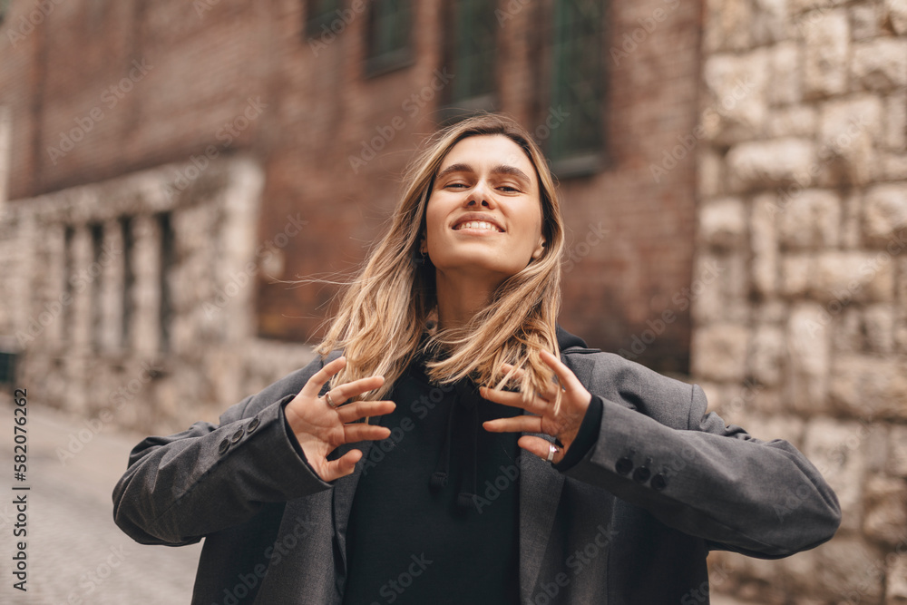 Trendy girl with blonde hairstyle posing outside. Woman in grey jacket and hoodie walking on the street outdoors. Tourist happy woman posing in the city. Optimistic lady walks outside and fix her hair Trendy girl with blonde hairstyle posing outside. Woman in grey jacket and hoodie walking on the street outdoors. Tourist happy woman posing in the city. Optimistic lady walks outside and fix her hair