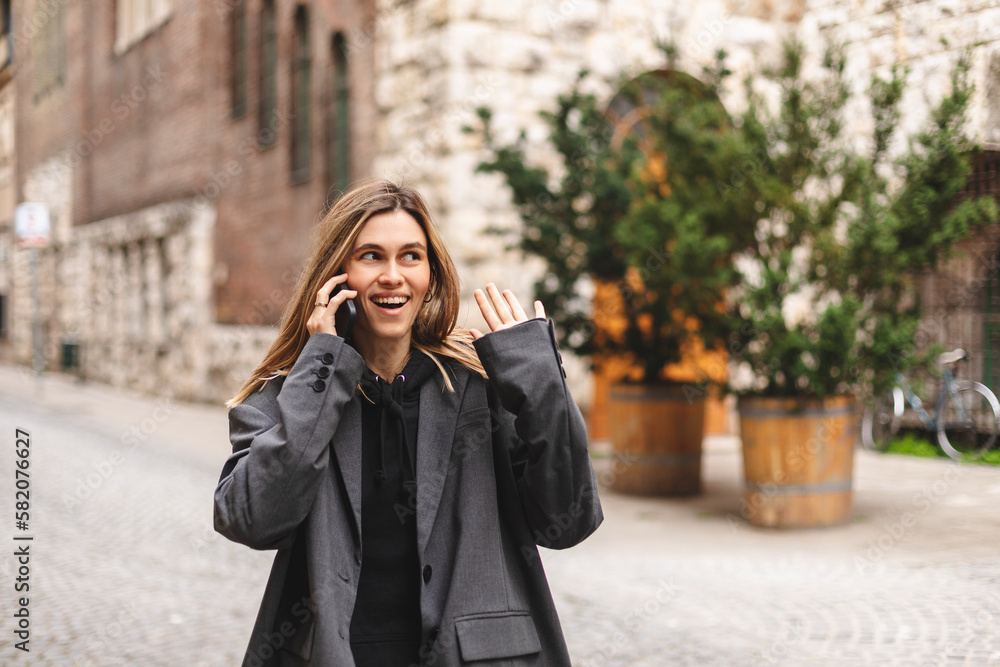 Young caucasian attractive smiling blonde woman in a grey jacket talking on a mobile phone flirting and twisting a lock of hair on her finger walking outside. Girl flirting. Young caucasian attractive smiling blonde woman in a grey jacket talking on a mobile phone flirting and twisting a lock of hair on her finger walking outside. Girl flirting.