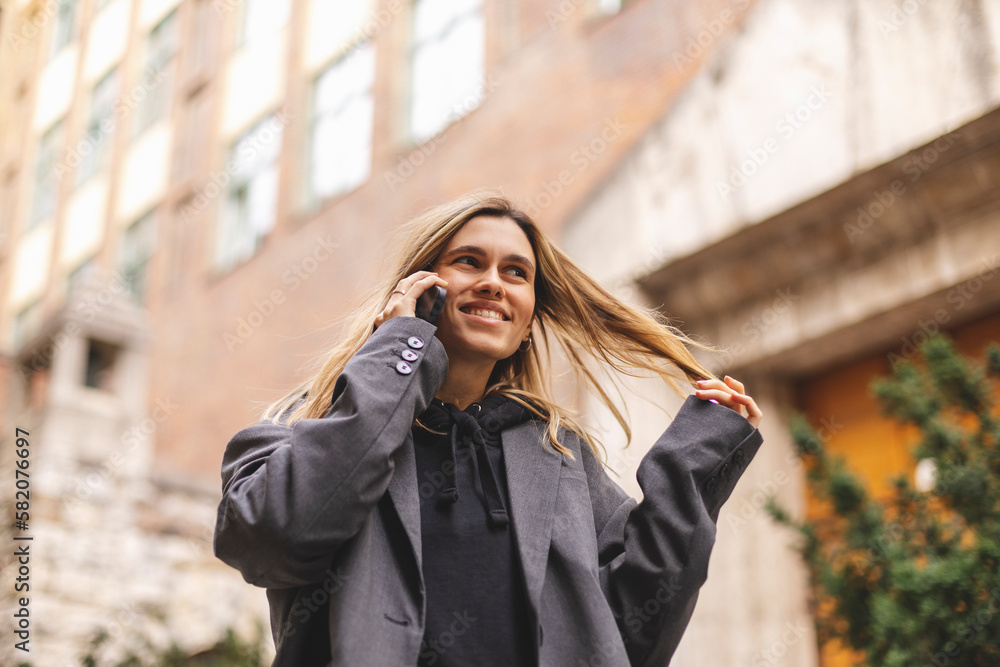 Surprised young caucasian attractive smiling blonde woman in a grey jacket talking on a mobile phone hear good news, open mouth and twisting a lock of hair on her finger walking outside. Girl flirting