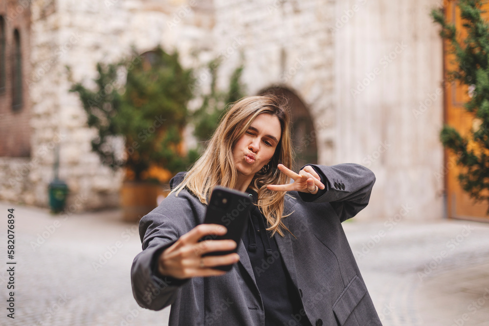 Fototapeta premium Happy young woman photographing herself using her mobile phone. Caucasian female taking selfie with her smart phone at city. Woman show peace sign and blow kiss, talk by video call, wear grey jacket.