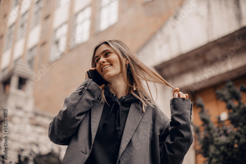 Surprised young caucasian attractive smiling blonde woman in a grey jacket talking on a mobile phone hear good news, open mouth and twisting a lock of hair on her finger walking outside. Girl flirting