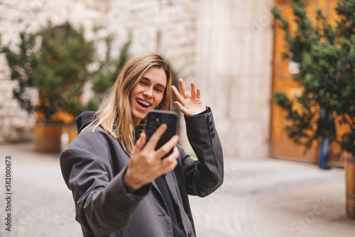 Woman waiving saying hello happy and smiling, friendly welcome gesture. Portrait of young beautiful attractive smiling cheerful girl taking selfie saying hello outside outdoors.