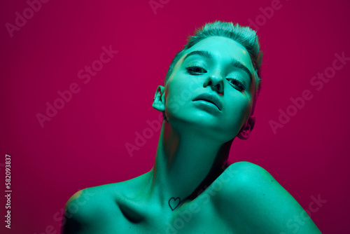 Fantasy. Beautiful young girl with short blonde hair posing with bare shoulders against pink studio background in green neon light. Cyberpunk style. Concept of futurism, digital world, robot, art