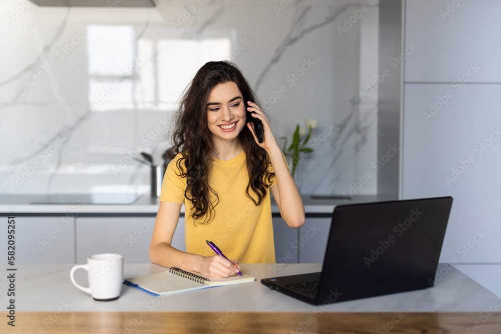 Portrait of a young woman talking the phone and working on laptop in kitchen Portrait of a young woman talking the phone and working on laptop in kitchen