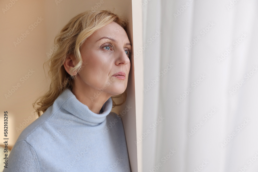 Upset middle aged woman near window at home. Loneliness concept Upset middle aged woman near window at home. Loneliness concept
