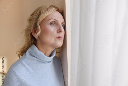 Upset middle aged woman near window at home. Loneliness concept