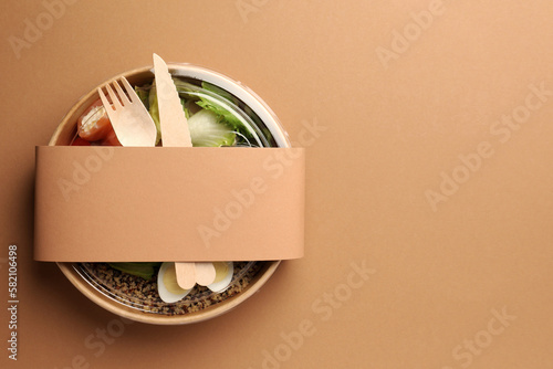 Tasty food in container with wooden fork and knife on beige background, top view. Space for text