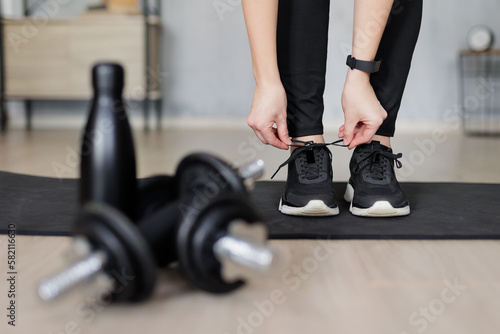 sport and healthy lifestyle concept - woman preparing for training at home and tying shoelaces on sneakers - dumbbells and bottle of water on first plan