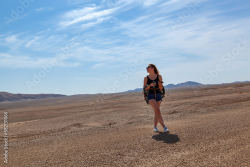 The girl is standing on the background of the desert in sunglasses and smiling. The model is wearing short shorts and a shirt. The girl enjoys a sunny day on vacation