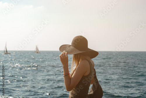 A girl in a black hat looks towards the sea. Two sailboats are sailing on the horizon. The model trims a hat in sunny weather on the seashore. The frame is illuminated by the sun