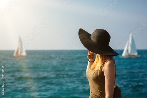 Silhouette of a girl on the background of the blue sea and blue sky. sailing yachts sail across the sea and the model looks in their direction. The black hat with blond hair visible under
