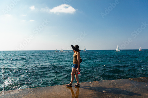 A girl in short shorts, a mask and a black hat stands on a pier by the sea. Yachts and sailboats can be seen on the Horikhont. Blue sea and blue sky on vacation