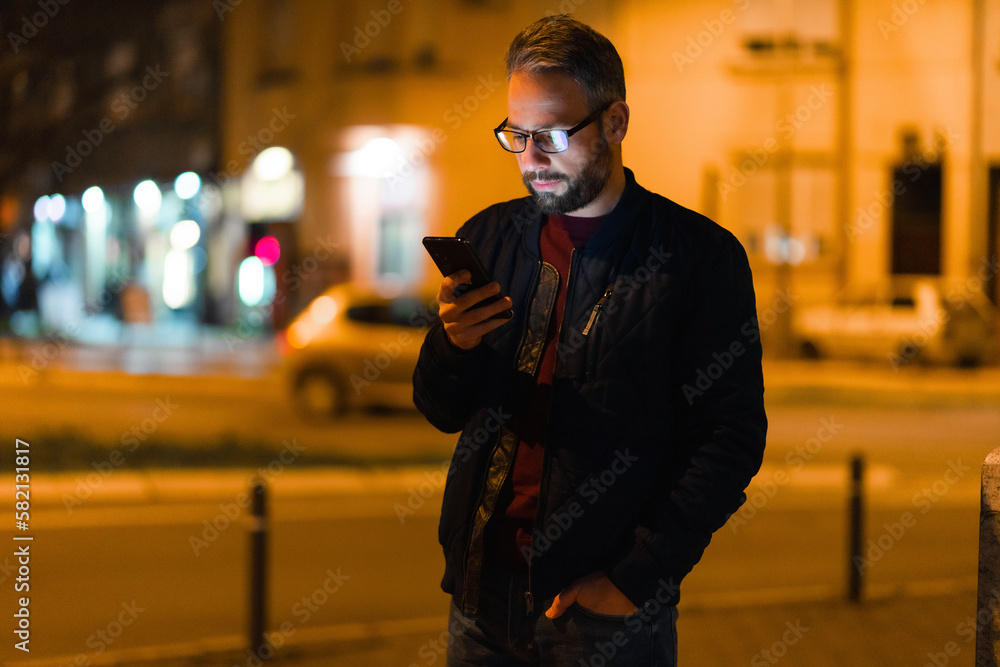 A man with glasses is walking at night in the city and using a smartphone. - obrazy, fototapety, plakaty 