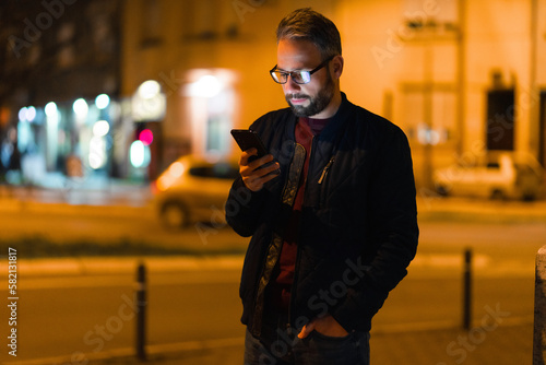 A man with glasses is walking at night in the city and using a smartphone. photo