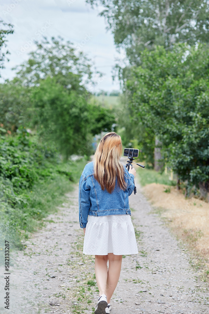 Elegant millennial woman broadcasting a video, using a tripod and phone, while walking outside in the summer park. Blogging lifestyle. Elegant millennial woman broadcasting a video, using a tripod and phone, while walking outside in the summer park. Blogging lifestyle.