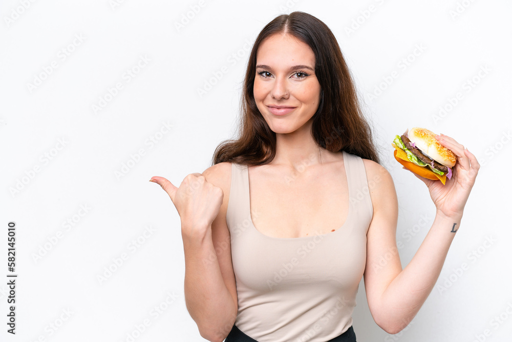 Young caucasian woman holding a burger isolated on white background pointing to the side to present a product