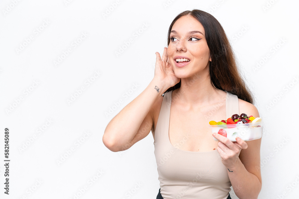 Young caucasian woman holding a bowl of fruit isolated on white background listening to something by putting hand on the ear Young caucasian woman holding a bowl of fruit isolated on white background listening to something by putting hand on the ear