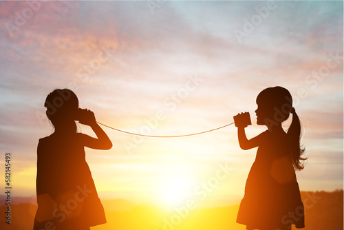 Silhouette of Two female kids stands and talking by using string can phone on beautiful sunset sky photo