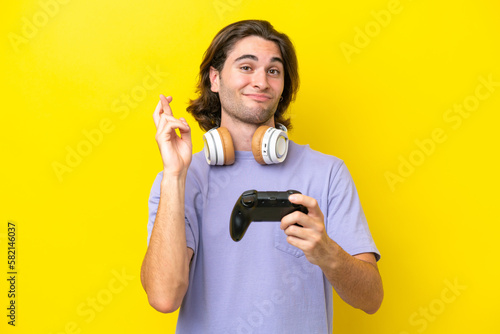 Young handsome caucasian man playing with a video game controller over isolated on yellow background with fingers crossing and wishing the best