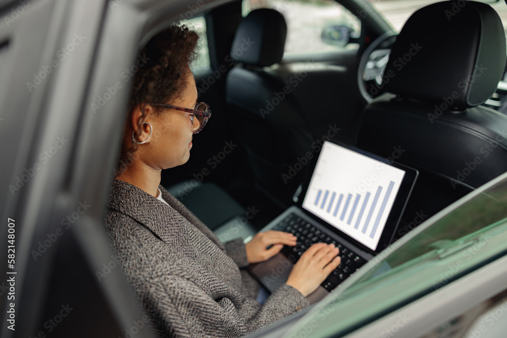Focused woman analyst working on laptop sitting car leather backseat on the way to office