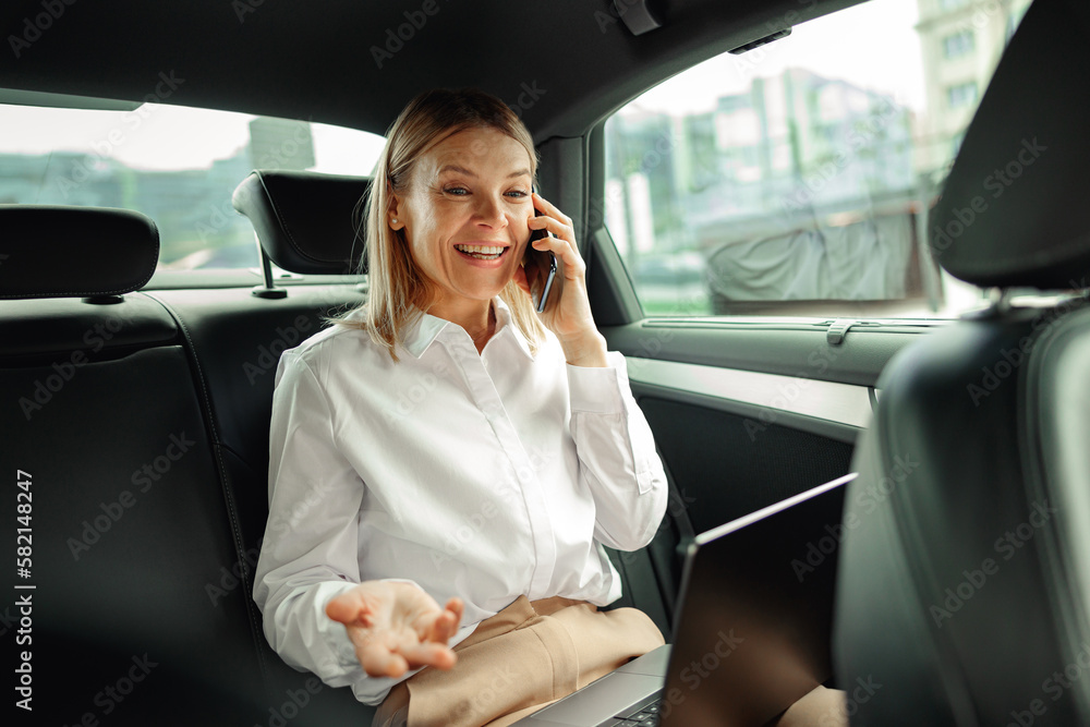 Female entrepreneur working on laptop and talking phone during traveling to office in luxury car