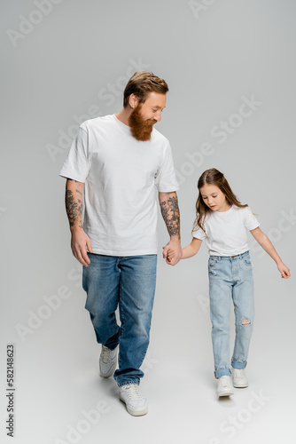 Tattooed man holding hand of daughter while walking on grey background.