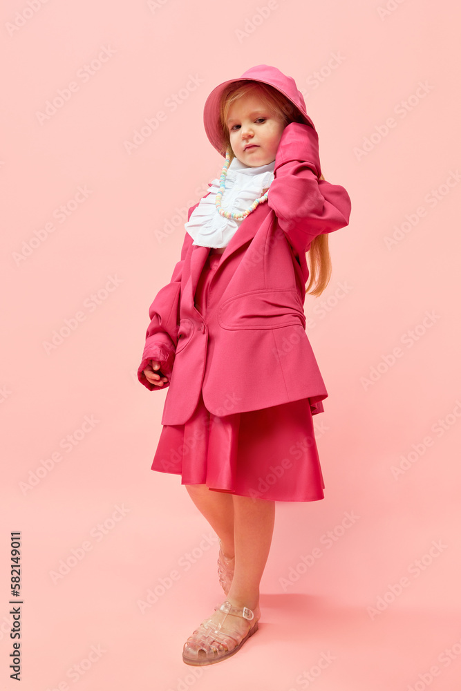 Photo of little, stylish girl touching headdress and wearing big pink, oversized clothes to imitate her mother over pink background. Full-length