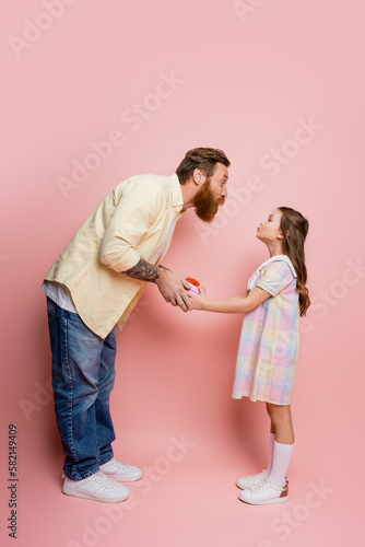 Side view of man pouting lips while giving gift box to daughter on pink background.