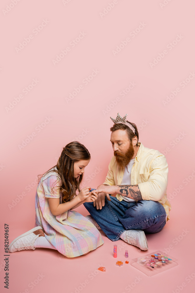 Preteen girl applying nail polish on hand of bearded father with crown headband on pink background. Preteen girl applying nail polish on hand of bearded father with crown headband on pink background.