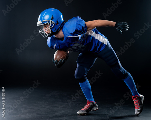 Full length portrait of a man in a blue american football uniform against a black background. Sportsman in a helmet with a ball. 