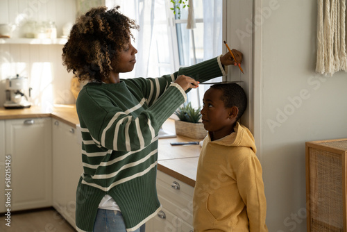 Fotografie Caring african american mother measures changes height of child son on wall with pencil at home