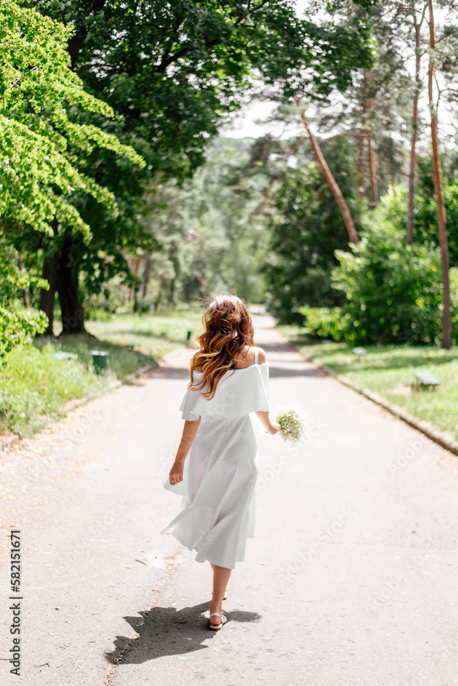 A slender beautiful girl in a white dress with a white bouquet in her hands walks cheerfully along the park path. Her dress and hair fly in the wind A slender beautiful girl in a white dress with a white bouquet in her hands walks cheerfully along the park path. Her dress and hair fly in the wind