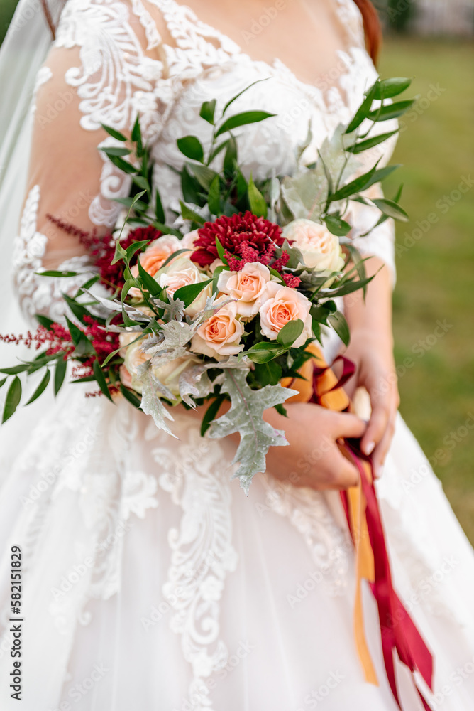 A bouquet of burgundy chrysanthemums and delicate pink roses, decorated with green leaves and red ribbons, in the hands of a bride in a white dress with lace, on a blurred background