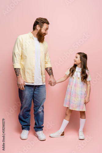 Side view of smiling tattooed man holding hand of preteen daughter on pink background.