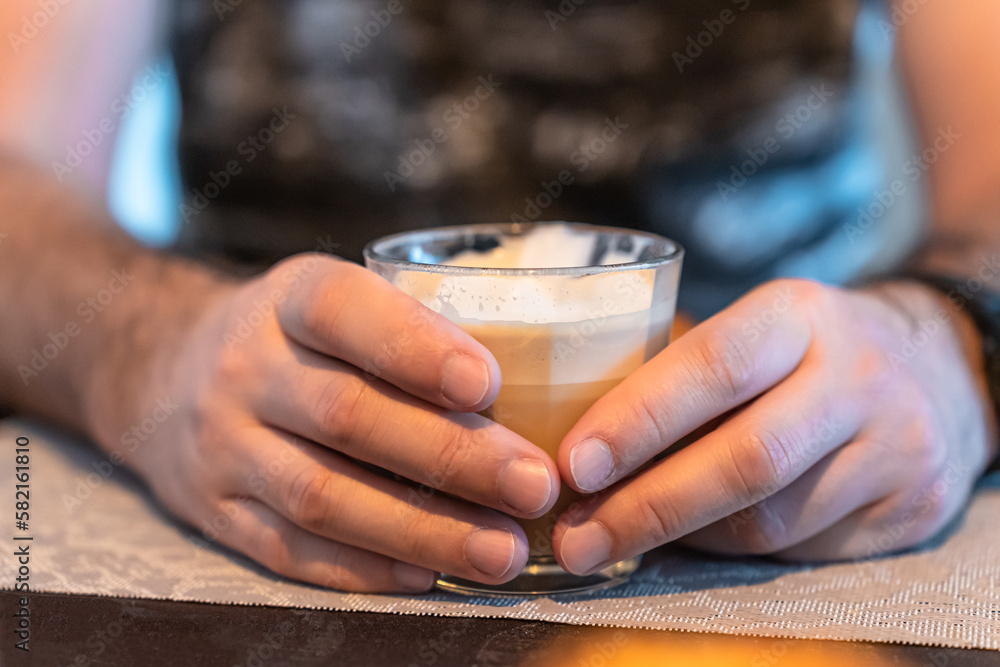 Focus in the foreground on the hands of a man who holds a glass of cappuccino. Focus in the foreground on the hands of a man who holds a glass of cappuccino.