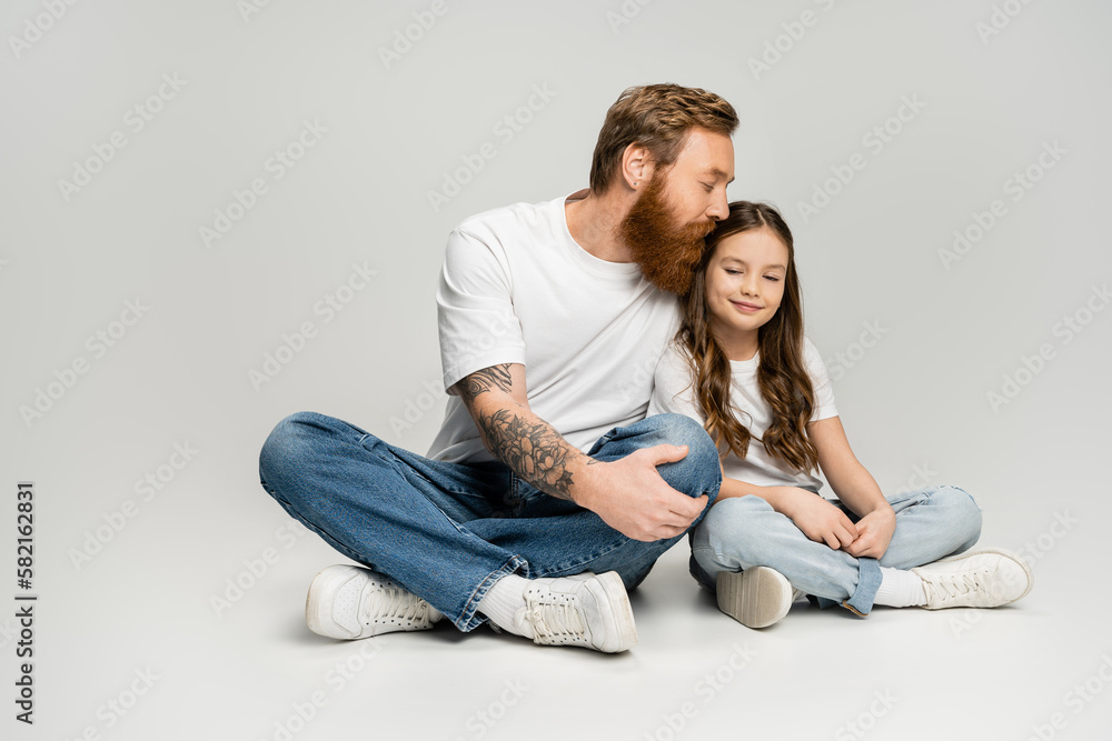Bearded man in jeans and t-shirt kissing smiling preteen daughter on grey background. Bearded man in jeans and t-shirt kissing smiling preteen daughter on grey background.