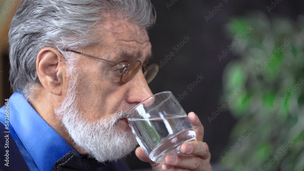 In the frame, a close-up of the face of the grandfather of an elderly man takes medicine, swallows a pill with water, in glasses with a gray beard In the frame, a close-up of the face of the grandfather of an elderly man takes medicine, swallows a pill with water, in glasses with a gray beard