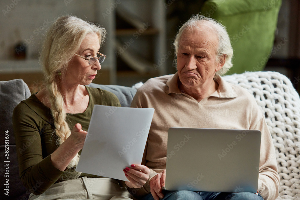 Serious mature people, man and woman sitting on sofa at home and working together with laptop and papers. Checking documents. Concept of family, relationship, lifestyle, love, retirement Serious mature people, man and woman sitting on sofa at home and working together with laptop and papers. Checking documents. Concept of family, relationship, lifestyle, love, retirement