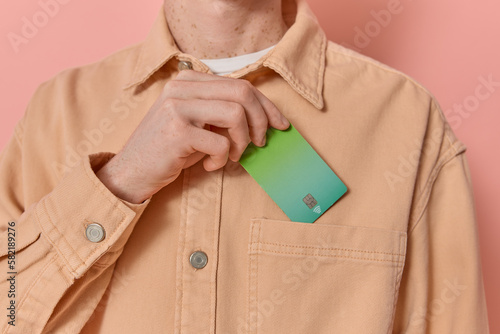 Anonymous man puts his green credit card in his pocket after payment on pink background copy space. Male consumer makes use of contactless payment methods like NFC. Modern banking, finance concept. 
