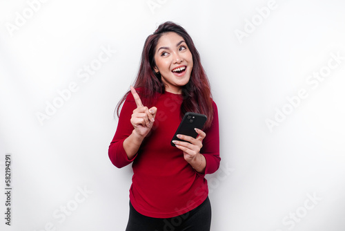 Excited Asian woman wearing red top pointing at the copy space on top of her while holding her phone, isolated by white background