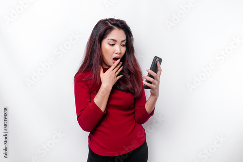 Shocked Asian woman wearing red top, holding her phone, isolated by white background