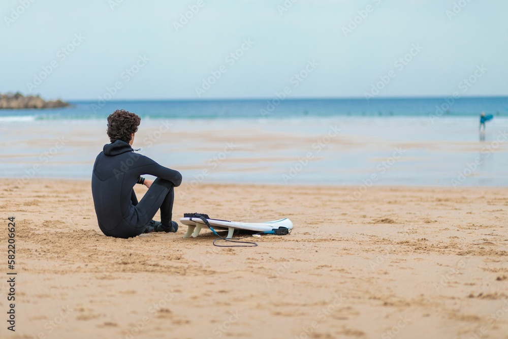 Curly-haired surfer sitting on the beach sand and looking at the seascape captured from behind
