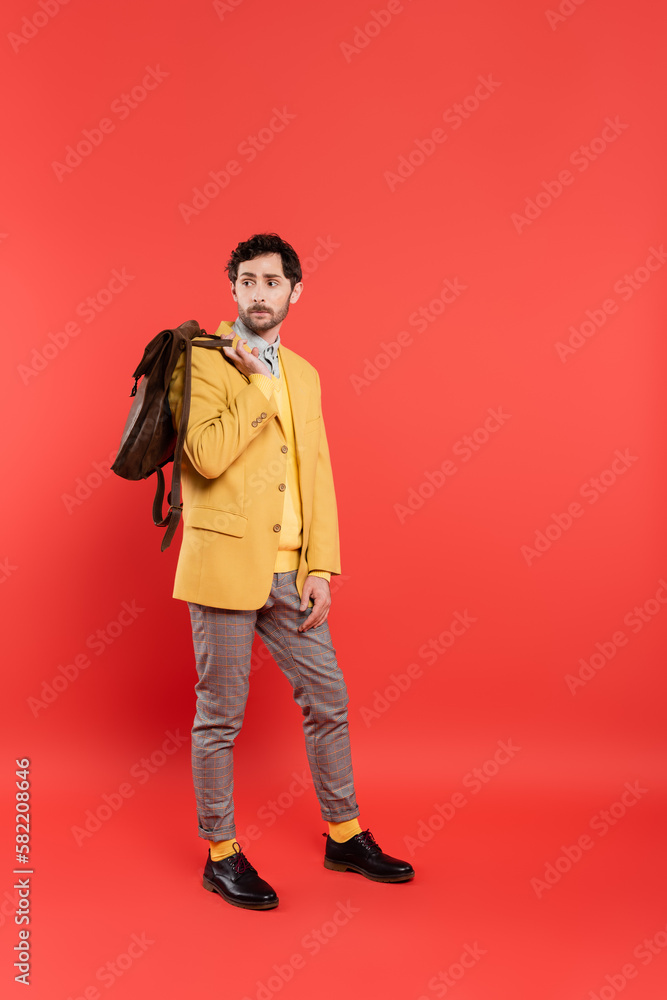 Full length of stylish model in yellow blazer holding backpack on coral red background. Full length of stylish model in yellow blazer holding backpack on coral red background.