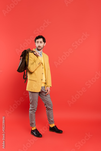 Full length of stylish model in yellow blazer holding backpack on coral red background.
